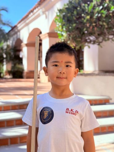 A boy holding a wooden stick, wearing a white B.K.D. Wushu t-shirt.