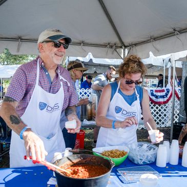 Two people serving food at an outdoor event under a tent.