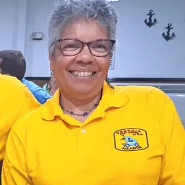 Smiling woman in a yellow shirt with a logo, wearing glasses and jewelry.
