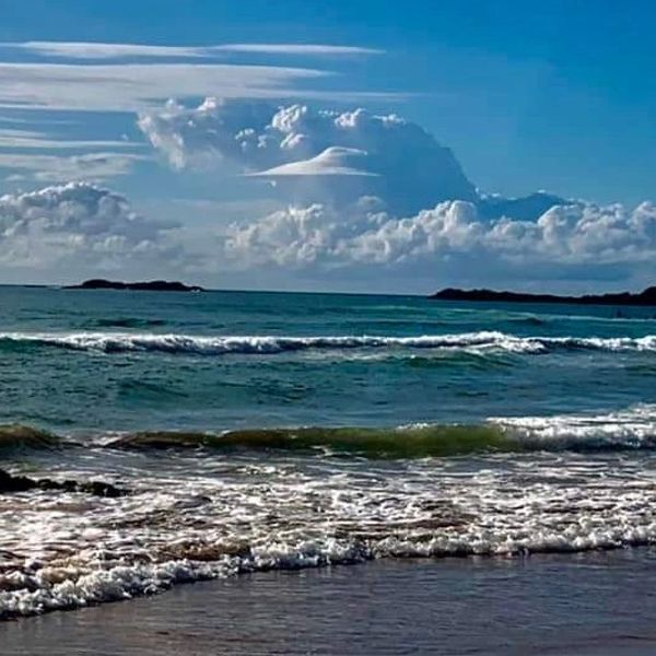 Ocean waves gently crash on the shore under a blue sky with striking cloud formations.