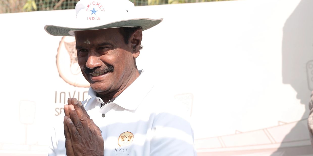 Man in white cricket attire greeting with folded hands and smiling.