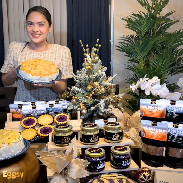 Woman proudly presenting a cheese cake surrounded by various packaged food products and festive decorations.