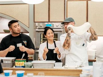Three chefs preparing food together in a kitchen, one stretching pizza dough.