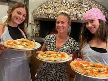 Three women proudly holding freshly made pizzas in front of a pizza oven.
