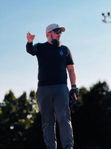 Man wearing a cap, sunglasses, and glove, gesturing outdoors against a blue sky.