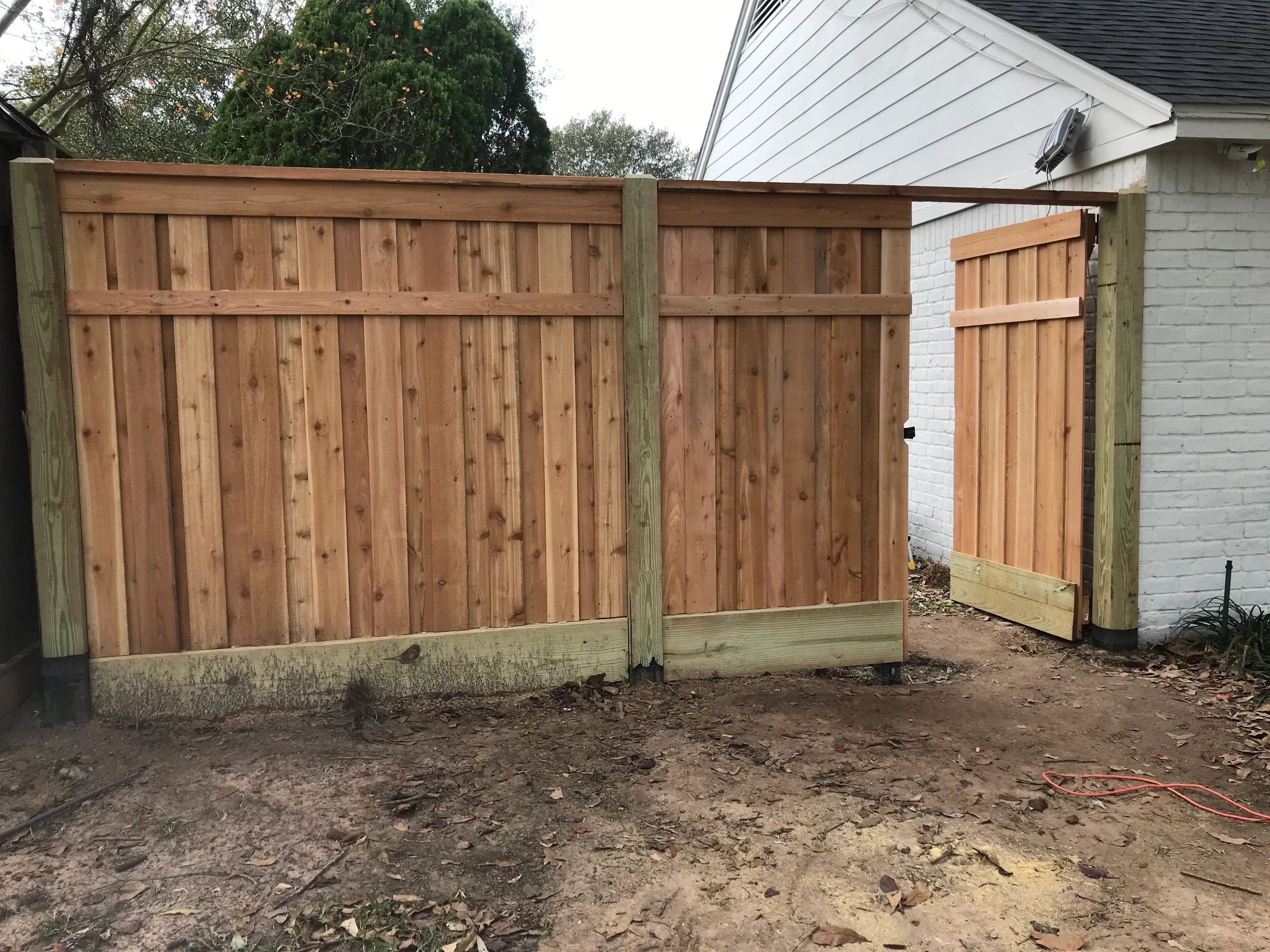 Newly installed wooden fence with an open gate beside a white brick house.