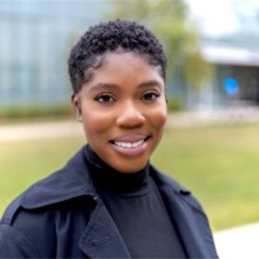 Black woman smiling in blue trench coat. Has rich skin with natural hair. 