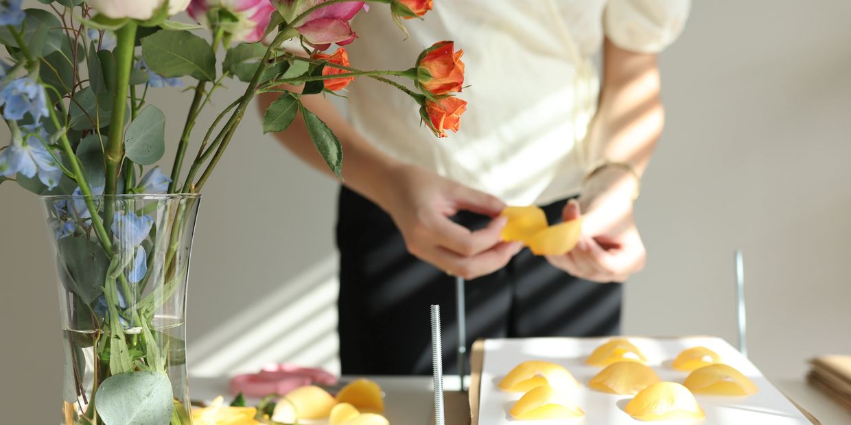 A table with a vase of flowers and an open flower press being filled with yellow rose petals.