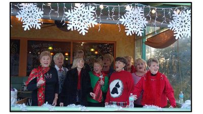 A group of elderly women singing together, festive decorations above them.