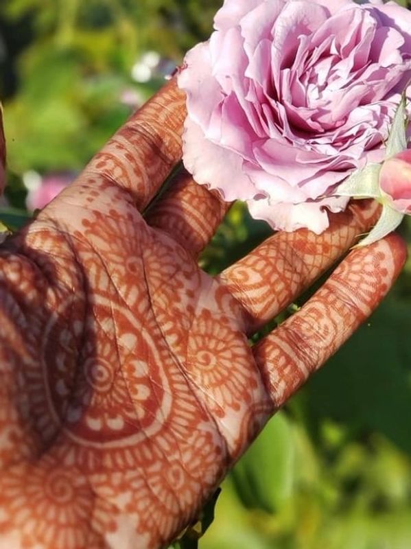 A hand with intricate henna designs holding a delicate pink rose.