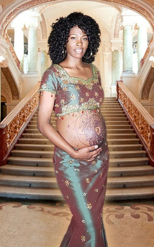 Pregnant woman in traditional attire with henna on belly, standing in a grand staircase hall.