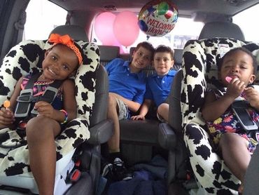 Four children in a car with welcome home balloons and two in cow-print car seats.