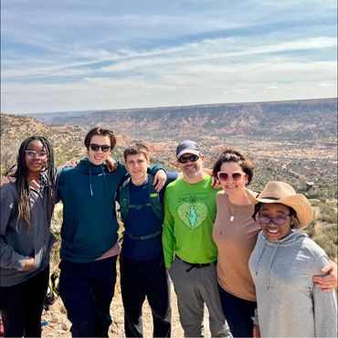 Group of six people smiling on a scenic hiking trail with mountains in the background.
