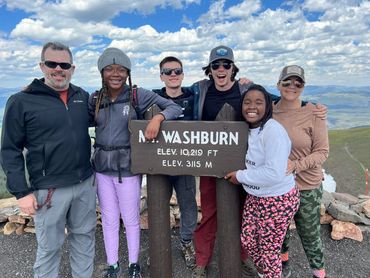 Group of six smiling hikers posing at Mt. Washburn summit sign.
