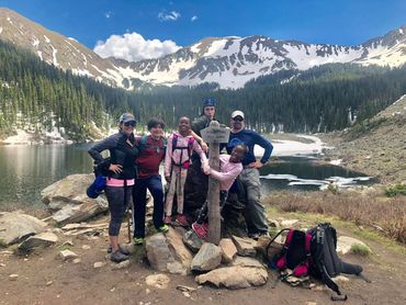 A happy group of hikers posing by Williams Lake sign with snowy mountains in the background.