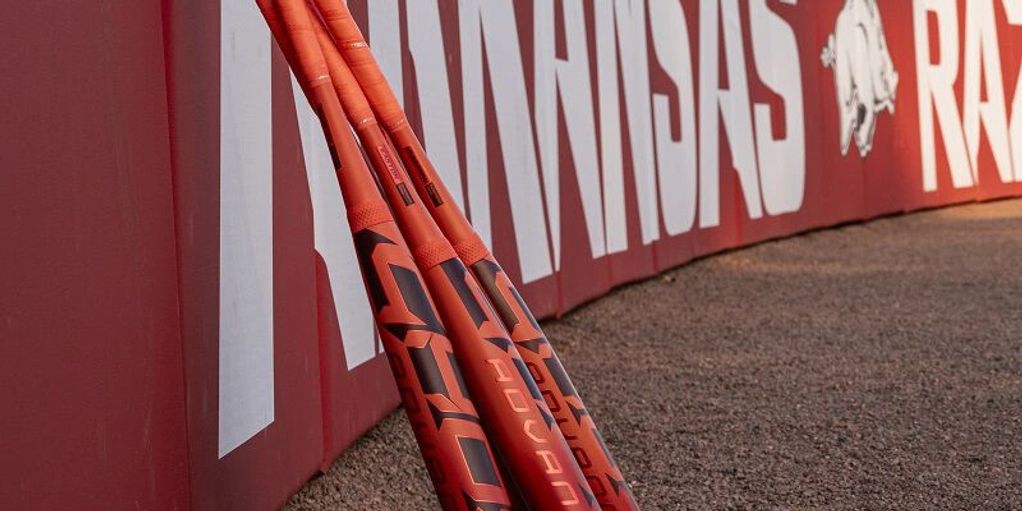 Three orange baseball bats leaning against a red Arkansas Razorbacks wall.