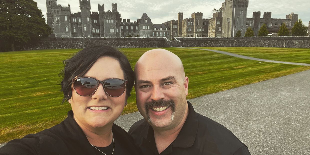 Smiling couple takes a selfie in front of a large historic castle.