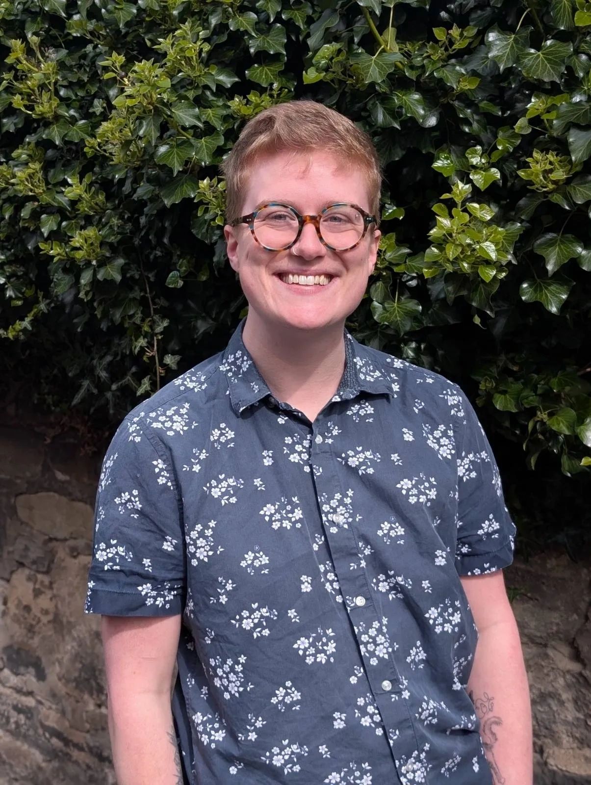 Smiling person with glasses and floral shirt standing outdoors.
