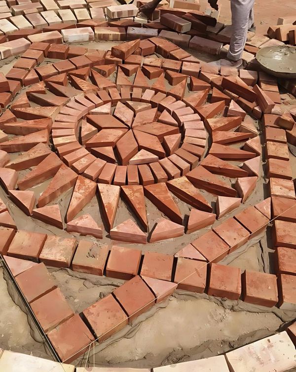 Intricate circular pattern of red bricks being laid on sand.