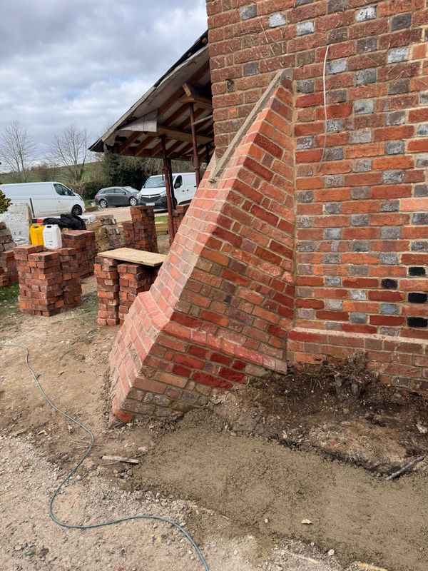 A leaning brick structure attached to a brick wall, under construction outdoors.