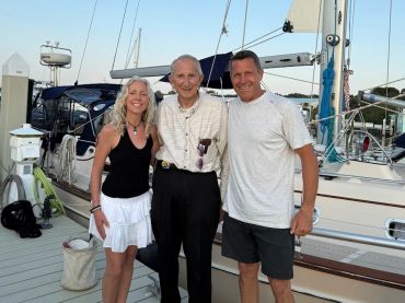 Three people smiling on a dock in front of a sailboat at sunset.