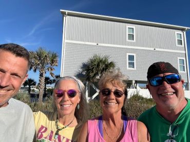 Four friends smiling outdoors with palm trees and a house in the background.