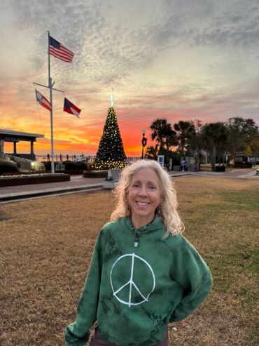 Smiling woman in green peace symbol sweatshirt at sunset near Christmas tree and flags.