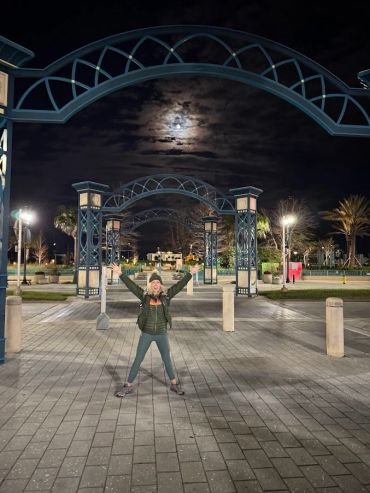 Person joyfully posing under illuminated arches at night with moon in the cloudy sky.