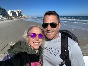 Couple smiling on a sunny beach with the ocean and buildings in the background.