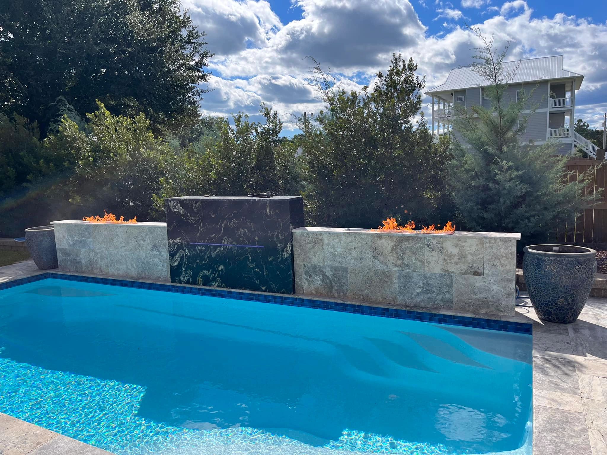 A modern pool with fire features and lush greenery under a partly cloudy sky.