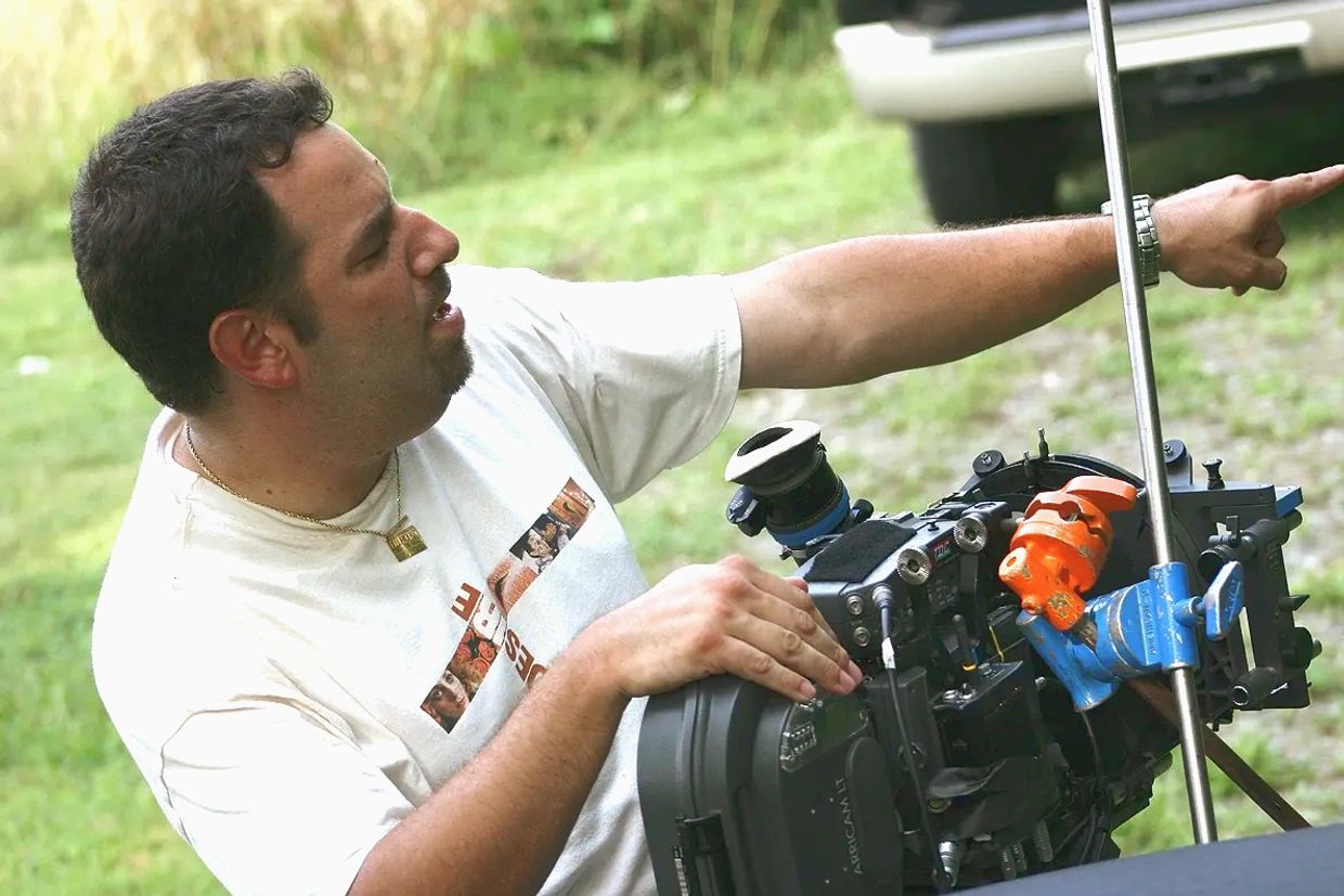 Man operating a professional film camera outdoors, pointing at something.