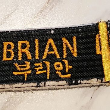 Worn black martial arts belt with "MASTER BRIAN" embroidered in yellow and rank stripes.