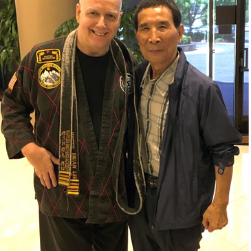 Two men posing together indoors, one in martial arts attire with a black belt.