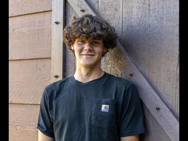 Young man with curly hair smiling, leaning against a wooden wall.