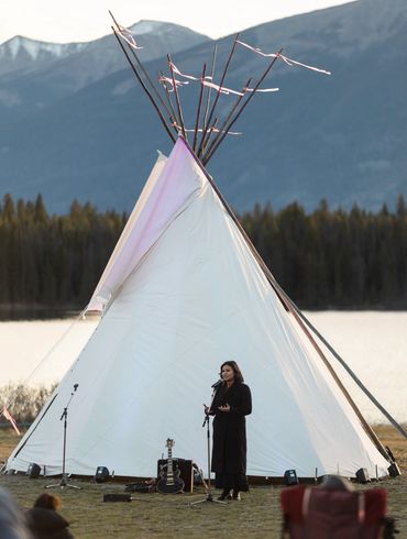 Woman speaking into a microphone in front of a large white teepee outdoors.