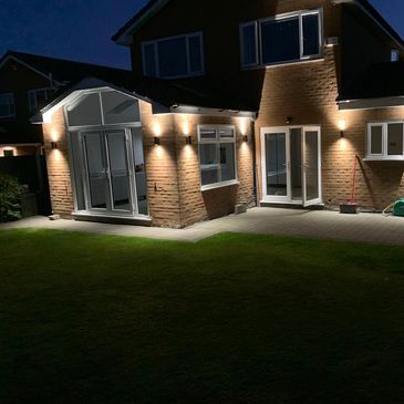A well-lit brick house exterior at night with a lawn and patio area.