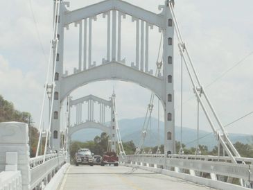 Vehicles crossing a white suspension bridge with mountains in the background.