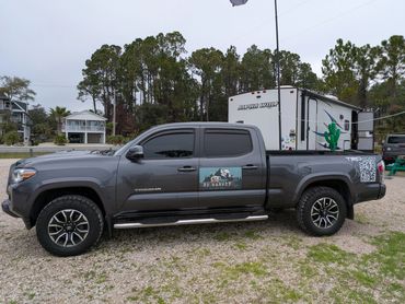 Gray Toyota Tacoma truck with RV Harvey logo and QR code on side, parked on gravel near RV.