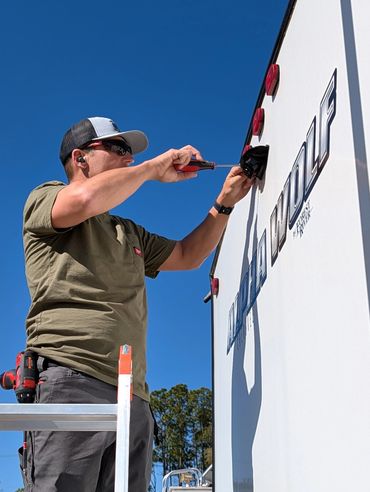 Man fixing a light on an Alpha Wolf RV under clear blue sky.