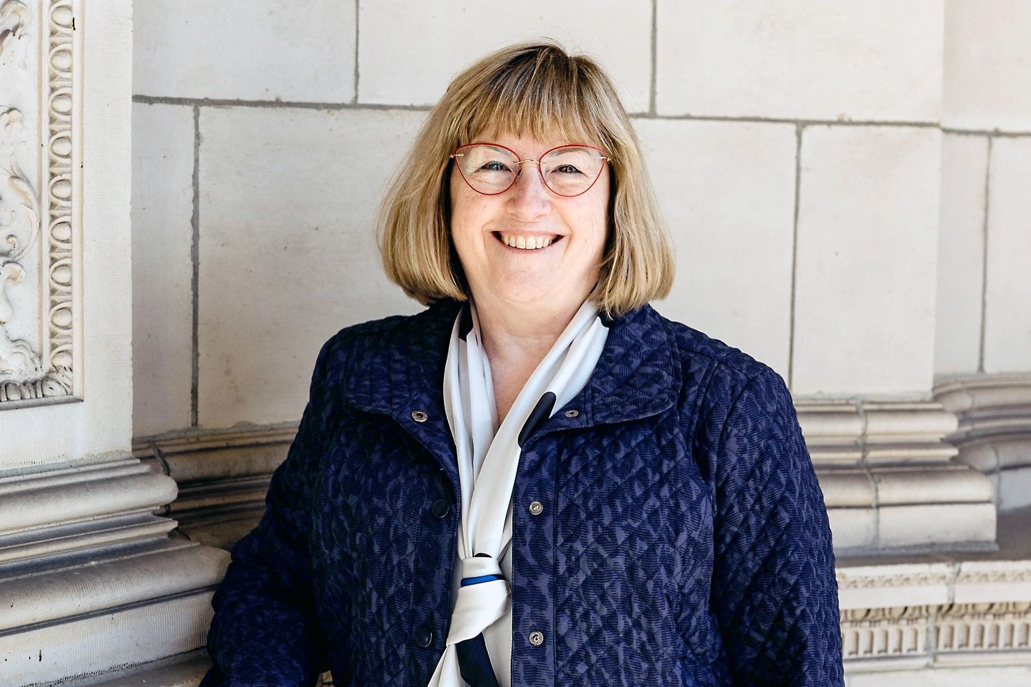 Smiling woman, financial consultant Susan Goodison, in blue jacket with glasses near a stone wall.