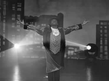 Performer in a dramatic pose on stage with cityscape backdrop and spotlight.
