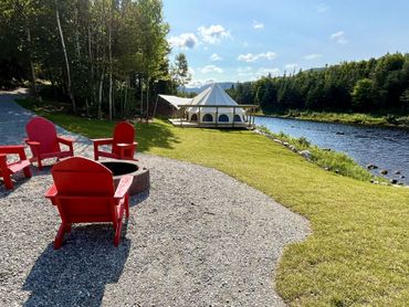 Red chairs around a fire pit by a river with a white tent and trees in the background.