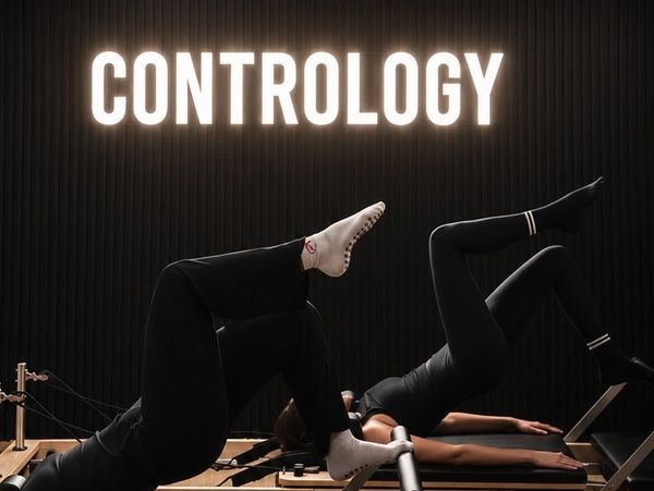 Two women exercising on pilates reformers under a bright 'CONTROL OLOGY' sign.