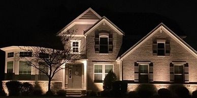 A well-lit house at night with shrubs and a tree.