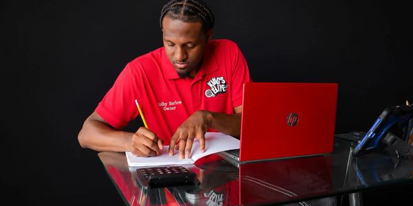 Man in red shirt working at a glass desk with a red laptop and calculator.