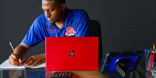 Man in blue shirt writing notes with a red HP laptop in front of him.