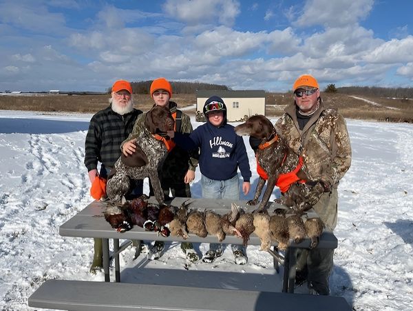 Group of hunters in orange gear holding harvested birds in an open field under clear blue sky.