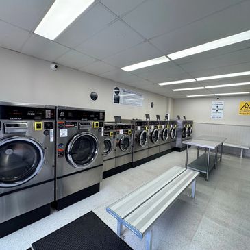 Modern laundromat with multiple stainless steel washers and folding benches.