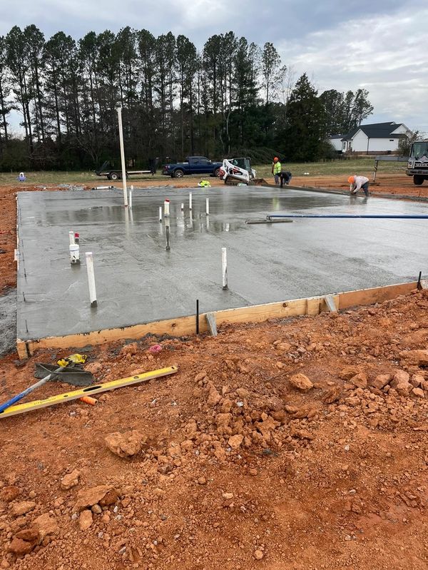 Workers finishing a freshly poured concrete foundation with pipes protruding.