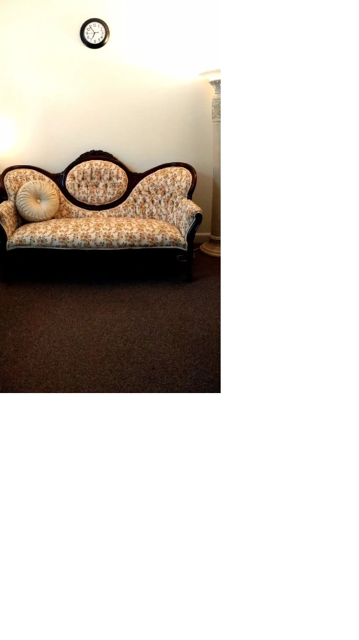 A grandma's sofa in well lit office with a clock above the sofa.  A brick and mortar office.